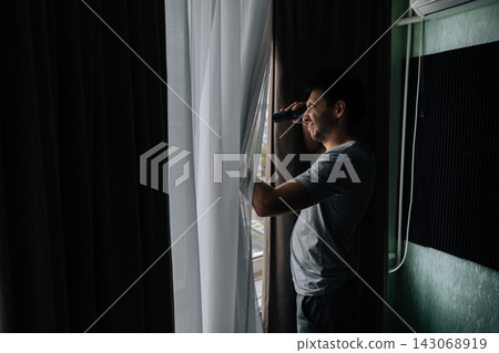 Remote view of man looking through monocular, subtly peeking between translucent and dark curtains, watching outside world with focused attention and sense of intrigue. Remote view of man looking through monocular, subtly peeking between translucent and dark curtains, watching outside world with focused attention and sense of intrigue. 143068919
