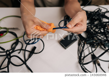 Close-up of person hands cleaning blue cable with an orange cloth, surrounded by a chaotic assortment of black and green audio, signal, and power cables on a white table. 143068946