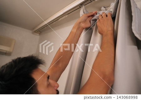 Close-up of man reaching up to attach light-colored curtain to white track rod on ceiling, focusing on setting up home decor in apartment, representing domestic life. Concept of household. Close-up of man reaching up to attach light-colored curtain to white track rod on ceiling, focusing on setting up home decor in apartment, representing domestic life. Concept of household. 143068998