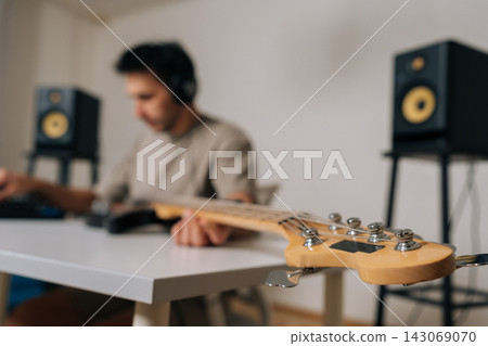 Selective focus of musician man wearing headphones and playing bass guitar, adjusting strings while connected to audio mixer for recording or home practice, focus on foreground. Selective focus of musician man wearing headphones and playing bass guitar, adjusting strings while connected to audio mixer for recording or home practice, focus on foreground. 143069070