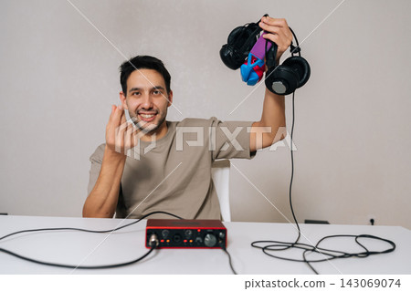 Happy man smiling and showing Korean finger heart gesture with one hand, holding professional headphones and audio interface, representing love for music production and sound engineering. Happy man smiling and showing Korean finger heart gesture with one hand, holding professional headphones and audio interface, representing love for music production and sound engineering. 143069074