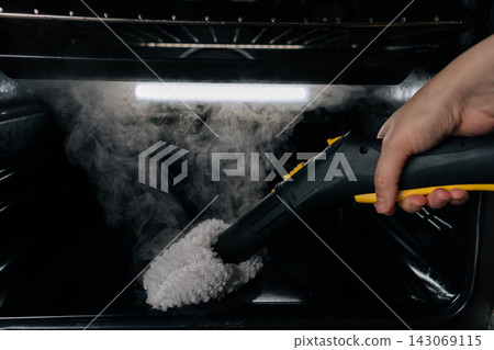Close-up view of housewife hand cleaning oven interior with steam cleaner, using hot vapor for effective and chemical-free domestic kitchen appliance disinfection and deep hygiene. Close-up view of housewife hand cleaning oven interior with steam cleaner, using hot vapor for effective and chemical-free domestic kitchen appliance disinfection and deep hygiene. 143069115
