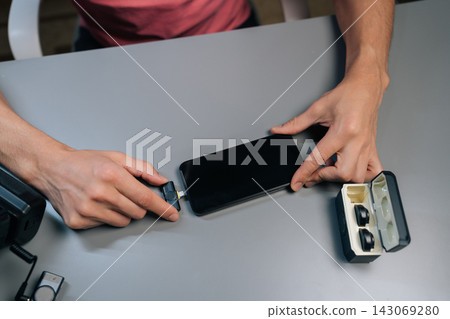Top view of content creator hands setting up professional audio recording system by plugging wireless lavalier microphone receiver with adapter into mobile phone for interviews. Top view of content creator hands setting up professional audio recording system by plugging wireless lavalier microphone receiver with adapter into mobile phone for interviews. 143069280