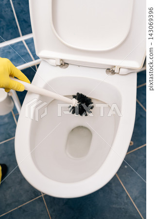Top view of person hand in yellow protective glove cleaning ceramic toilet bowl with brush, maintaining cleanliness and hygiene standards in residential or commercial washroom environment. Top view of person hand in yellow protective glove cleaning ceramic toilet bowl with brush, maintaining cleanliness and hygiene standards in residential or commercial washroom environment. 143069396