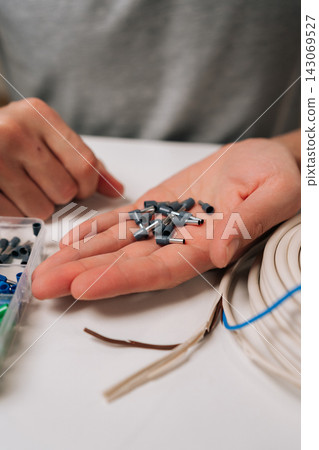 Vertical close-up view of hands selecting wire ferrules from plastic organizer next to coiled electrical cable on white table, depicting diy, home improvement and professional electrical work. 143069527