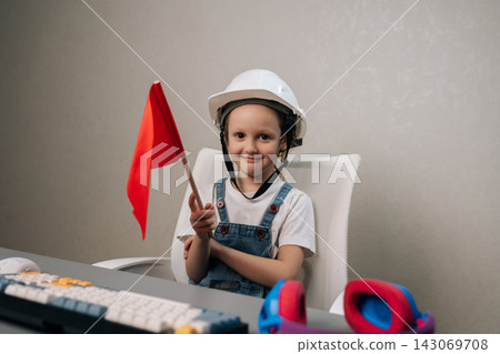 Young child wearing white safety helmet and blue overalls, smiling looking at camera while holding red warning flag at desk with keyboard, symbolizing safety and future careers. Young child wearing white safety helmet and blue overalls, smiling looking at camera while holding red warning flag at desk with keyboard, symbolizing safety and future careers. 143069708