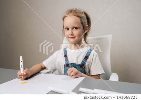 Studio portrait of young girl smiling and holding marker, creating art on white sheet of paper while sitting at grey desk, fostering creativity and childhood development. Studio portrait of young girl smiling and holding marker, creating art on white sheet of paper while sitting at grey desk, fostering creativity and childhood development. 143069723
