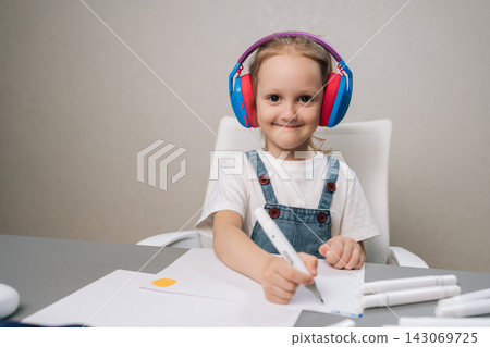 Smiling young child wearing colorful headphones, sitting at desk and drawing on paper with marker, actively engaged in creative and educational activity. Smiling young child wearing colorful headphones, sitting at desk and drawing on paper with marker, actively engaged in creative and educational activity. 143069725