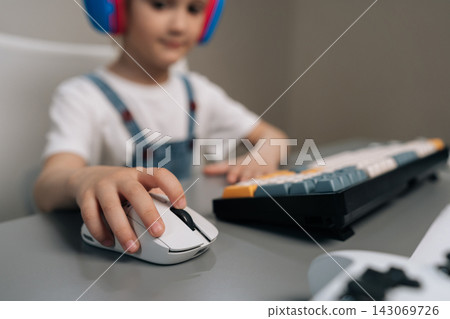Close-up of childs hand using computer mouse. Young gamer in headphones plays video games at home. Shallow depth of field focuses on modern equipment. Concept of esports, hobby, digital lifestyle. 143069726