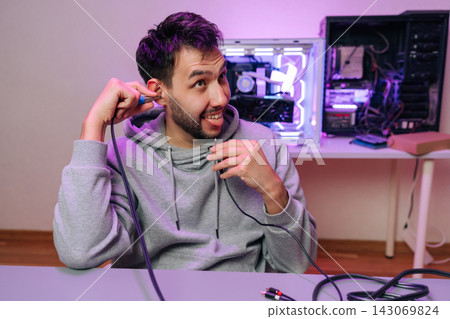 Young bearded man playfully sticks an audio jack to ear making silly face beside gaming pc setup, casual hoodie and cables scattered on desk. Young bearded man playfully sticks an audio jack to ear making silly face beside gaming pc setup, casual hoodie and cables scattered on desk. 143069824