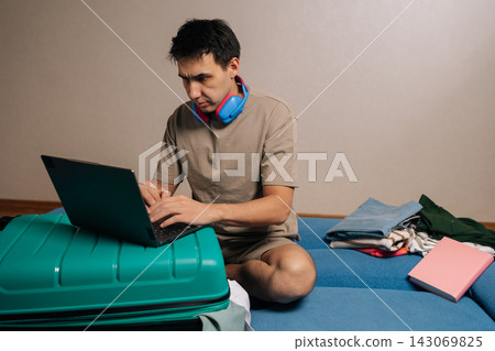 Portrait of traveler male preparing for travel, working on his laptop placed on a teal luggage, with neatly folded clothes stacked beside him on a couch, organizing logistics for his vacation. Portrait of traveler male preparing for travel, working on his laptop placed on a teal luggage, with neatly folded clothes stacked beside him on a couch, organizing logistics for his vacation. 143069825