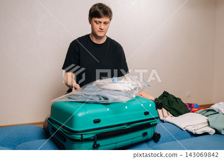 Portrait of traveler male compressing clothes into vacuum bag, optimizing space within a turquoise suitcase while preparing for a vacation, trip, or relocation, demonstrating smart packing techniques. Portrait of traveler male compressing clothes into vacuum bag, optimizing space within a turquoise suitcase while preparing for a vacation, trip, or relocation, demonstrating smart packing techniques. 143069842