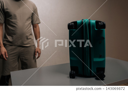 Person standing behind teal suitcase on a light background, representing travel preparation, packing a bag for a journey, anticipating departure, and the concept of going away. Person standing behind teal suitcase on a light background, representing travel preparation, packing a bag for a journey, anticipating departure, and the concept of going away. 143069872