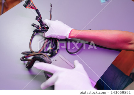 Technician hands in white gloves holding computer power supply wires. IT specialist repairing or building PC hardware on grey desk. Workspace illuminated by vibrant purple light. Technology concept. Technician hands in white gloves holding computer power supply wires. IT specialist repairing or building PC hardware on grey desk. Workspace illuminated by vibrant purple light. Technology concept. 143069891