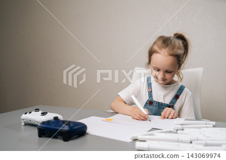 Focused young girl in overalls sitting at table drawing with markers. Kid ignores video game controllers, choosing creative hobby over digital entertainment. Soft light, neutral colors, copy space. Focused young girl in overalls sitting at table drawing with markers. Kid ignores video game controllers, choosing creative hobby over digital entertainment. Soft light, neutral colors, copy space. 143069974