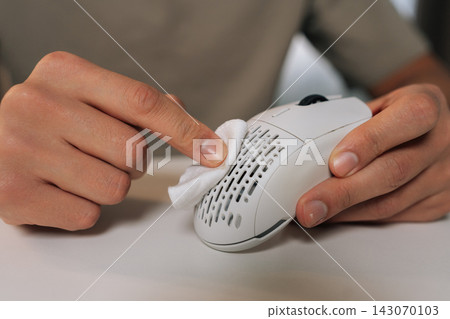 Person hands cleaning white wireless ergonomic computer mouse with small cleaning cloth, ensuring hygiene and proper maintenance of modern technology peripheral. Person hands cleaning white wireless ergonomic computer mouse with small cleaning cloth, ensuring hygiene and proper maintenance of modern technology peripheral. 143070103