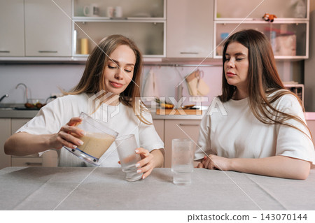 Two young women in kitchen. One pours homemade fruit smoothie from blender into glasses. Friend watches with skeptical expression. Concept for healthy lifestyle, diet, friendship, morning routine. Two young women in kitchen. One pours homemade fruit smoothie from blender into glasses. Friend watches with skeptical expression. Concept for healthy lifestyle, diet, friendship, morning routine. 143070144