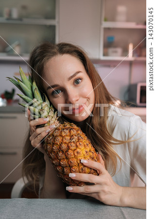 Portrait of happy young woman with blue eyes holding fresh pineapple. She smiles gently looking at camera in her kitchen. Healthy lifestyle, natural beauty and vegetarian diet concept. Portrait of happy young woman with blue eyes holding fresh pineapple. She smiles gently looking at camera in her kitchen. Healthy lifestyle, natural beauty and vegetarian diet concept. 143070145
