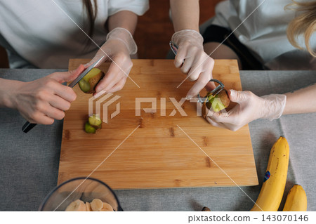 Top view of two people preparing healthy fruit salad together. Hands in disposable gloves peel kiwi with knife and peeler on wooden board. Concept of cooking, diet, and hygiene. Top view of two people preparing healthy fruit salad together. Hands in disposable gloves peel kiwi with knife and peeler on wooden board. Concept of cooking, diet, and hygiene. 143070146