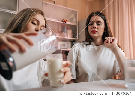 Two young women friends sit at table in kitchen. One pours milk from blender into glass for breakfast. Casual, domestic scene with soft lighting. Good for lifestyle, food, or relationship concepts. Two young women friends sit at table in kitchen. One pours milk from blender into glass for breakfast. Casual, domestic scene with soft lighting. Good for lifestyle, food, or relationship concepts. 143070164