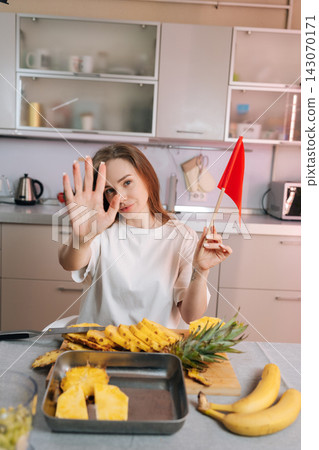 Young woman in kitchen holds red flag and shows stop hand gesture. Sliced pineapple and bananas are on table. Concept for dating red flags, boundaries, warnings, or healthy eating limits. Young woman in kitchen holds red flag and shows stop hand gesture. Sliced pineapple and bananas are on table. Concept for dating red flags, boundaries, warnings, or healthy eating limits. 143070171