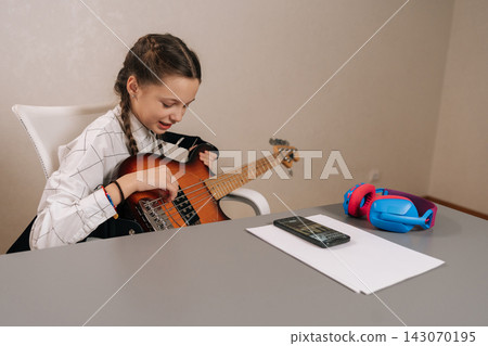 Young girl with pigtails focused on playing bass guitar, following online lesson on smartphone, developing new musical skills and a creative hobby at home. Young girl with pigtails focused on playing bass guitar, following online lesson on smartphone, developing new musical skills and a creative hobby at home. 143070195