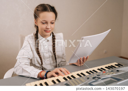 Focused pretty smiling young girl learning to play a musical instrument, focusing on her sheet music while practicing on an electronic keyboard, developing new skills and creativity. Focused pretty smiling young girl learning to play a musical instrument, focusing on her sheet music while practicing on an electronic keyboard, developing new skills and creativity. 143070197