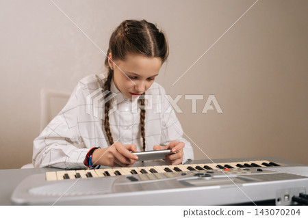 Focused preteen girl looking to screen, holding smartphone and sitting at synthesizer, using modern technology for music education and practicing piano or keyboard skills. Focused preteen girl looking to screen, holding smartphone and sitting at synthesizer, using modern technology for music education and practicing piano or keyboard skills. 143070204