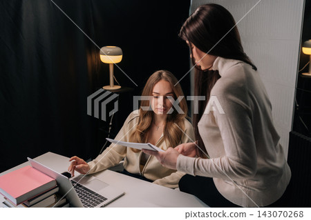 Two businesswomen collaborating in dark office setting, reviewing paper documents and providing advice during meeting, showing teamwork and professional interaction. Two businesswomen collaborating in dark office setting, reviewing paper documents and providing advice during meeting, showing teamwork and professional interaction. 143070268