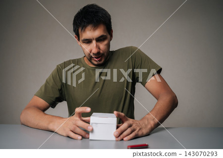 Portrait of young man unpacking new electronic cigarette using small knife sitting at table on isolated background. Concept of modern alternative to traditional smoking. Studio shot, copy space. Portrait of young man unpacking new electronic cigarette using small knife sitting at table on isolated background. Concept of modern alternative to traditional smoking. Studio shot, copy space. 143070293