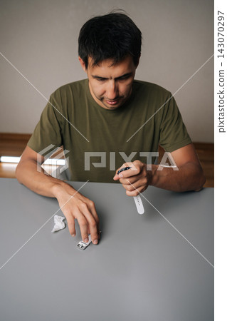 Vertical portrait of focused man assembling white strap onto smart watch, showcasing process of customizing or repairing wearable device sitting at table. Man connecting white smartwatch strap Vertical portrait of focused man assembling white strap onto smart watch, showcasing process of customizing or repairing wearable device sitting at table. Man connecting white smartwatch strap 143070297