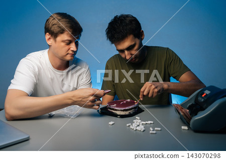 Portrait of two young men sitting at table eating cake laced with drugs, pills scattered on table near money counter, suggesting illegal activity and celebration of ill-gotten gains. Portrait of two young men sitting at table eating cake laced with drugs, pills scattered on table near money counter, suggesting illegal activity and celebration of ill-gotten gains. 143070298