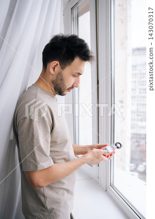 Vertical portrait of man measuring natural light near window with digital luxmeter, testing illumination levels for optimal home brightness, energy efficiency and comfortable indoor daylighting. Vertical portrait of man measuring natural light near window with digital luxmeter, testing illumination levels for optimal home brightness, energy efficiency and comfortable indoor daylighting. 143070317
