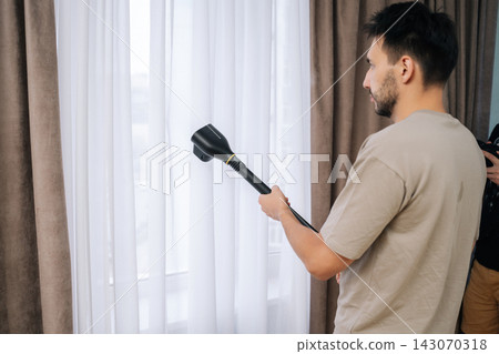 Homemaker male using handheld steam cleaner to sanitize and refresh sheer white curtains in modern home, ensuring deep cleaning and eliminating dust from fabric for improved air quality. Homemaker male using handheld steam cleaner to sanitize and refresh sheer white curtains in modern home, ensuring deep cleaning and eliminating dust from fabric for improved air quality. 143070318