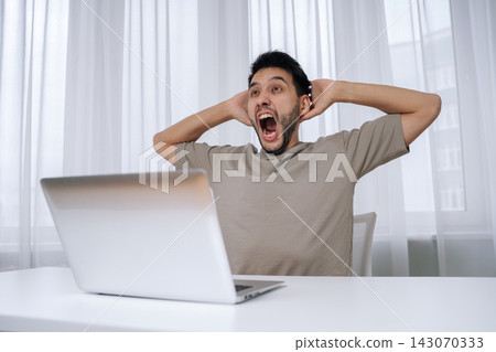 Young shouting man holding head hands with open mouth, experiencing shock and disbelief while looking at up laptop screen, reacting to unexpected bad news or critical mistake during remote work. Young shouting man holding head hands with open mouth, experiencing shock and disbelief while looking at up laptop screen, reacting to unexpected bad news or critical mistake during remote work. 143070333