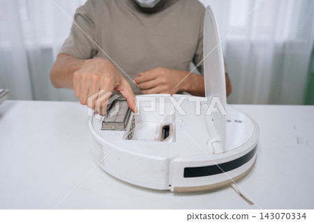 Person performing routine maintenance on robot vacuum cleaner, removing dirty filter to ensure efficient floor cleaning and smart home appliance longevity, close-up cropped shot. Person performing routine maintenance on robot vacuum cleaner, removing dirty filter to ensure efficient floor cleaning and smart home appliance longevity, close-up cropped shot. 143070334