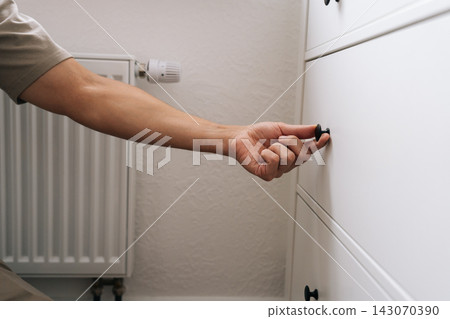 Close-up view of male hand pulling black knob to open white cabinet drawer. Minimalist home interior with radiator in background. Concept of daily routine, organization, and domestic life. Close-up view of male hand pulling black knob to open white cabinet drawer. Minimalist home interior with radiator in background. Concept of daily routine, organization, and domestic life. 143070390