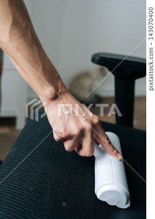 Close-up of hand using white lint roller to clean pet fur and dust from black fabric office chair. Domestic chore and household cleaning concept with cat blurred in background. Everyday routine. Close-up of hand using white lint roller to clean pet fur and dust from black fabric office chair. Domestic chore and household cleaning concept with cat blurred in background. Everyday routine. 143070400