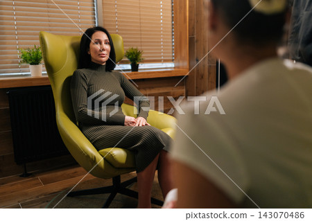 Female client seated in armchair in private therapy session, engaging with therapist, discussing mental health, challenges and progress in calm, confidential counseling setting 143070486