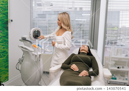 Cosmetic professional doctor preparing ultrasound facial lifting treatment settings on modern aesthetic machine for client relaxing in chair, focusing on skincare and beauty procedures in spa salon. 143070541