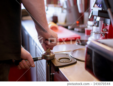 Making caffeine drink. Tamping coffee. Male barista hand pressing grinded beans powder in metal horn preparing aroma beverage in cafeteria. Making caffeine drink. Tamping coffee. Male barista hand pressing grinded beans powder in metal horn preparing aroma beverage in cafeteria. 143070544