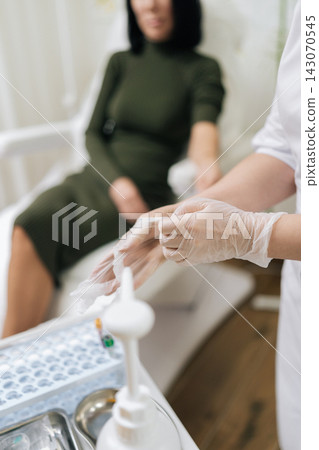 Medical worker pulling on sterile examination glove, ensuring safety and hygiene before medical procedure with blurred patient receiving care in background, close-up. Concept of healthy lifestyle. Medical worker pulling on sterile examination glove, ensuring safety and hygiene before medical procedure with blurred patient receiving care in background, close-up. Concept of healthy lifestyle. 143070545