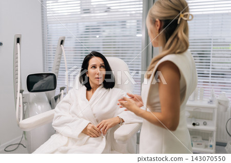 Side view of cosmetologist discussing treatment options with client during a professional consultation at modern beauty clinic, providing expert skincare advice and aesthetic services. Side view of cosmetologist discussing treatment options with client during a professional consultation at modern beauty clinic, providing expert skincare advice and aesthetic services. 143070550