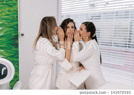 Three young adult doctors in white uniform sharing secret, whispering gossip and shocking news to colleague covering her mouth in disbelief, creating atmosphere of intrigue and communication. Three young adult doctors in white uniform sharing secret, whispering gossip and shocking news to colleague covering her mouth in disbelief, creating atmosphere of intrigue and communication. 143070593