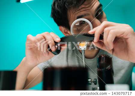 Young man using magnifying loupe to inspect roll of photographic film negative, analyzing images against bright teal background, engaging in analog hobby. Young man using magnifying loupe to inspect roll of photographic film negative, analyzing images against bright teal background, engaging in analog hobby. 143070717