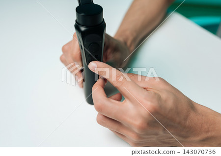 Close-up hand of unrecognizable blogger adjusting settings on black handheld electronic gimbal stabilizer, pressing buttons on device for steady video recording on clean, bright, isolated background. 143070736