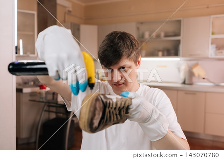 Young man in white t shirt wearing gloves, spraying and treating dirty sneaker on kitchen countertop, focused on cleaning, maintenance and hygiene. 143070780
