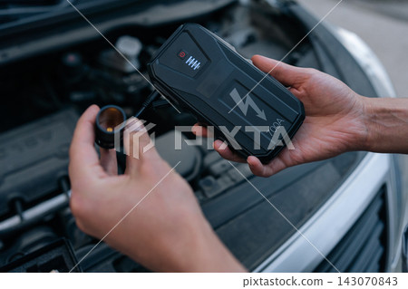 Close-up of hands holding portable car jump starter. Device ready for use with open engine bay in background. Concept for roadside assistance, vehicle maintenanceautomotive technology. Close-up of hands holding portable car jump starter. Device ready for use with open engine bay in background. Concept for roadside assistance, vehicle maintenanceautomotive technology. 143070843
