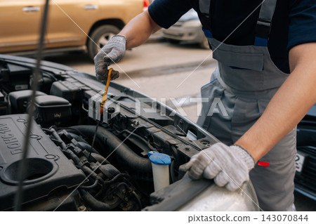 Close-up of mechanic hands in gloves checking car engine oil level with dipstick. Professional auto service and vehicle maintenance. Open hood at repair shop. Close-up of mechanic hands in gloves checking car engine oil level with dipstick. Professional auto service and vehicle maintenance. Open hood at repair shop. 143070844