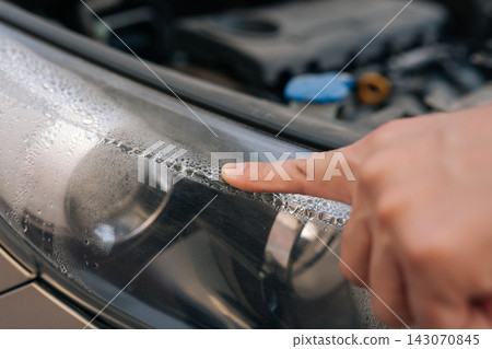Person finger inspecting water condensation forming inside modern car headlight, indicating potential repair or maintenance issue in vehicle. Person finger inspecting water condensation forming inside modern car headlight, indicating potential repair or maintenance issue in vehicle. 143070845